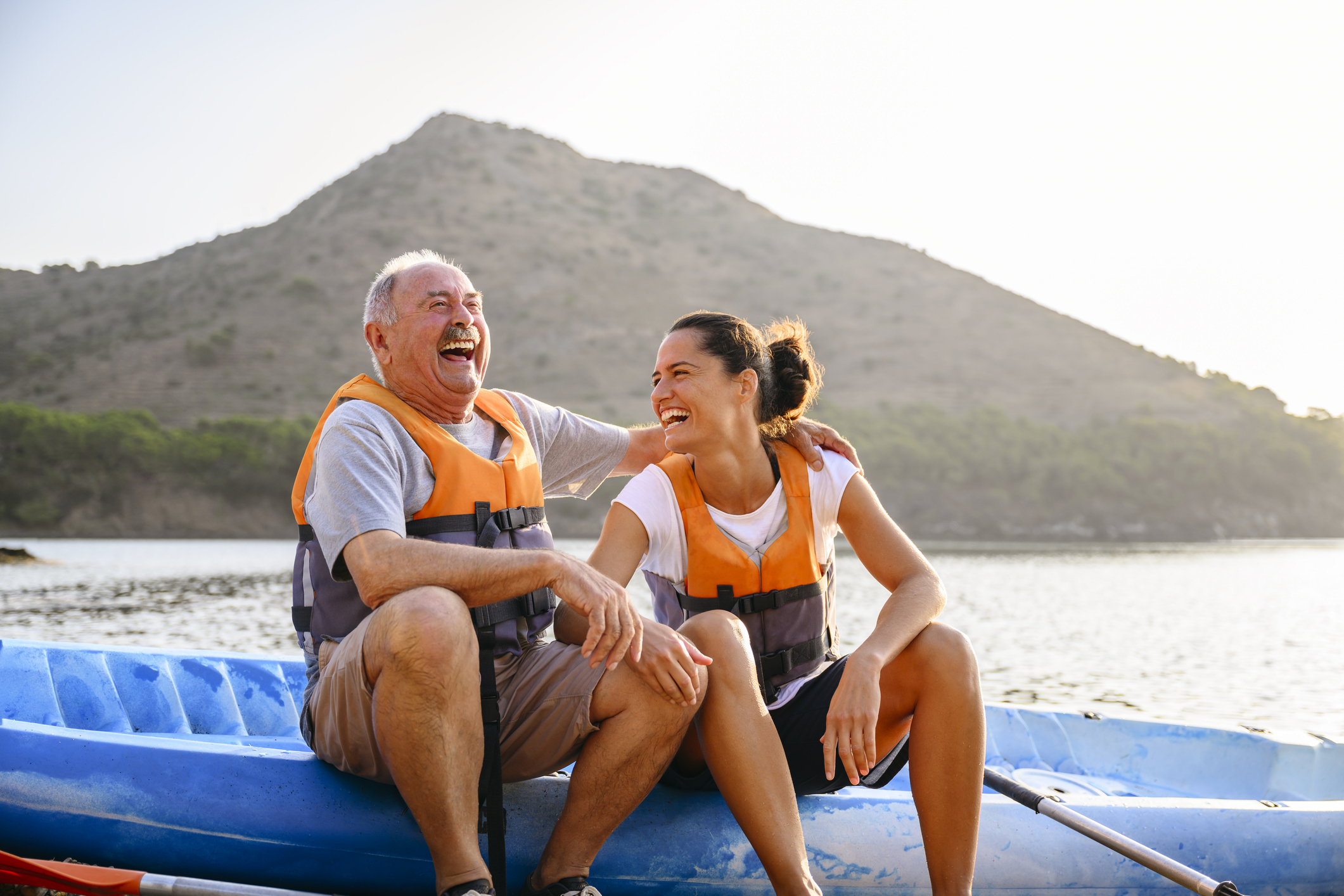 An older man and young woman with safety vests smiling and laughing while sitting on a blue kayak